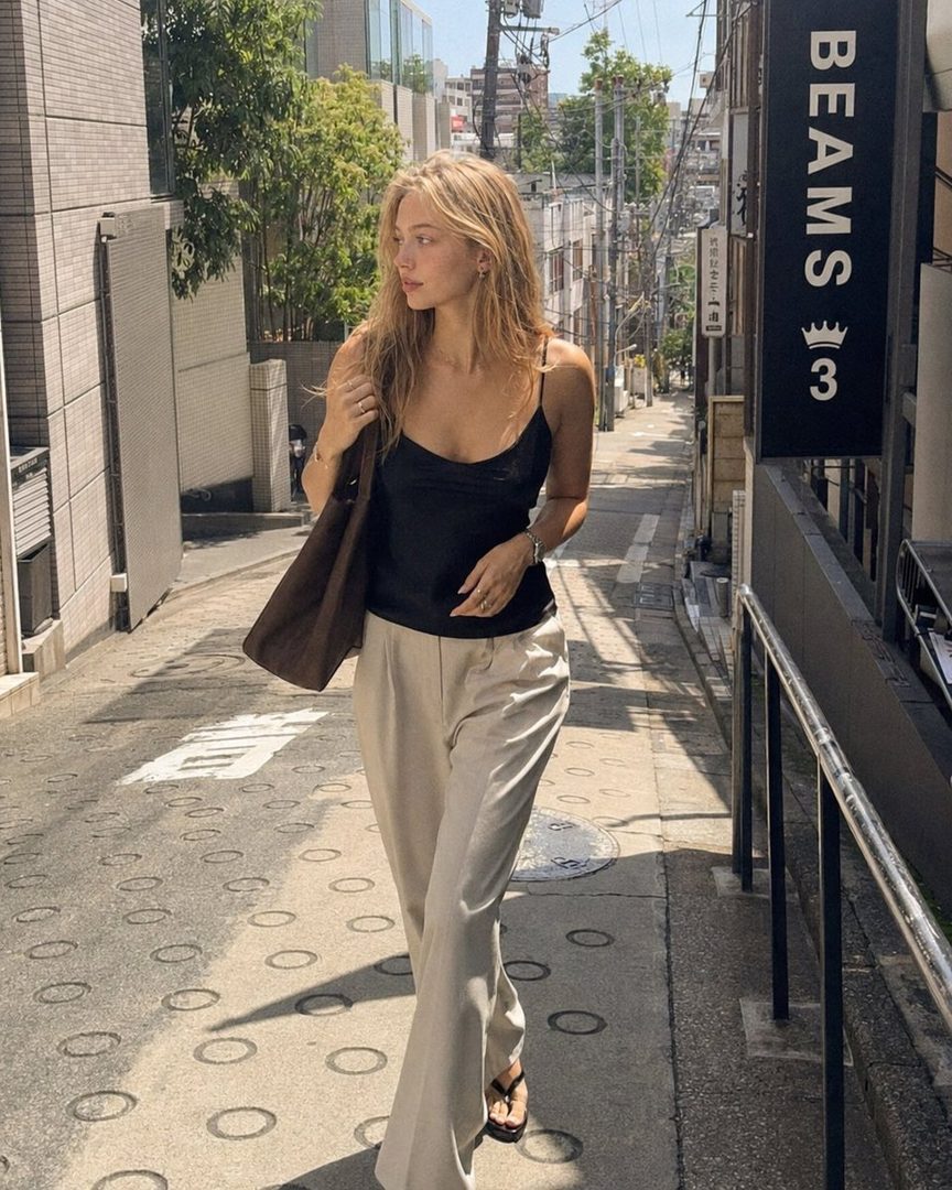 A young woman with blonde hair walks down a narrow city street on a sunny day, wearing a black camisole and beige wide-leg pants.