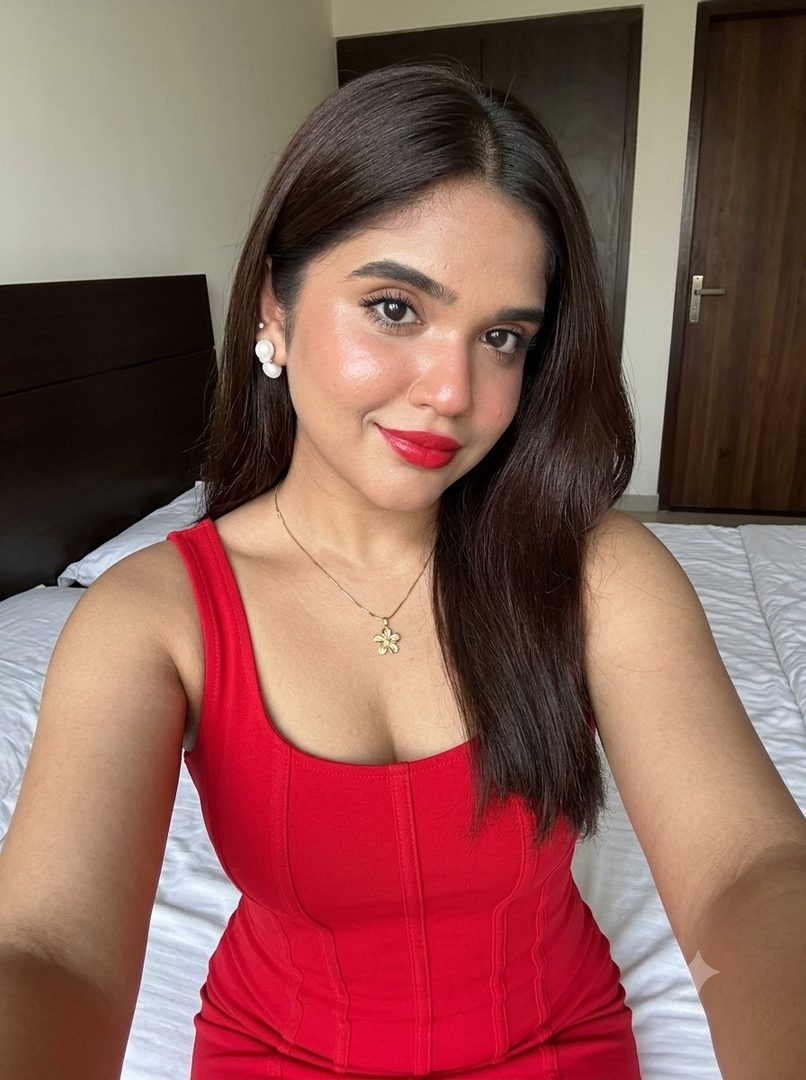 A young woman with long dark brown hair, wearing a red dress and red lipstick, takes a selfie in a bedroom.