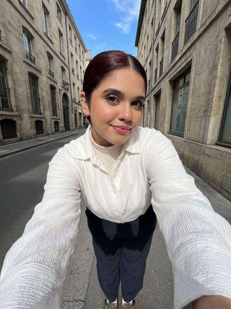A young woman with dark hair pulled back, wearing a white collared shirt and dark pants, takes a selfie on a European street with old buildings.