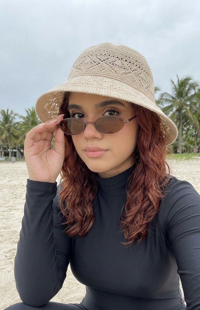 A young woman with long wavy brown hair, wearing a black long-sleeved swimsuit, a beige bucket hat, and sunglasses, takes a selfie on a sandy beach with palm trees in the background.