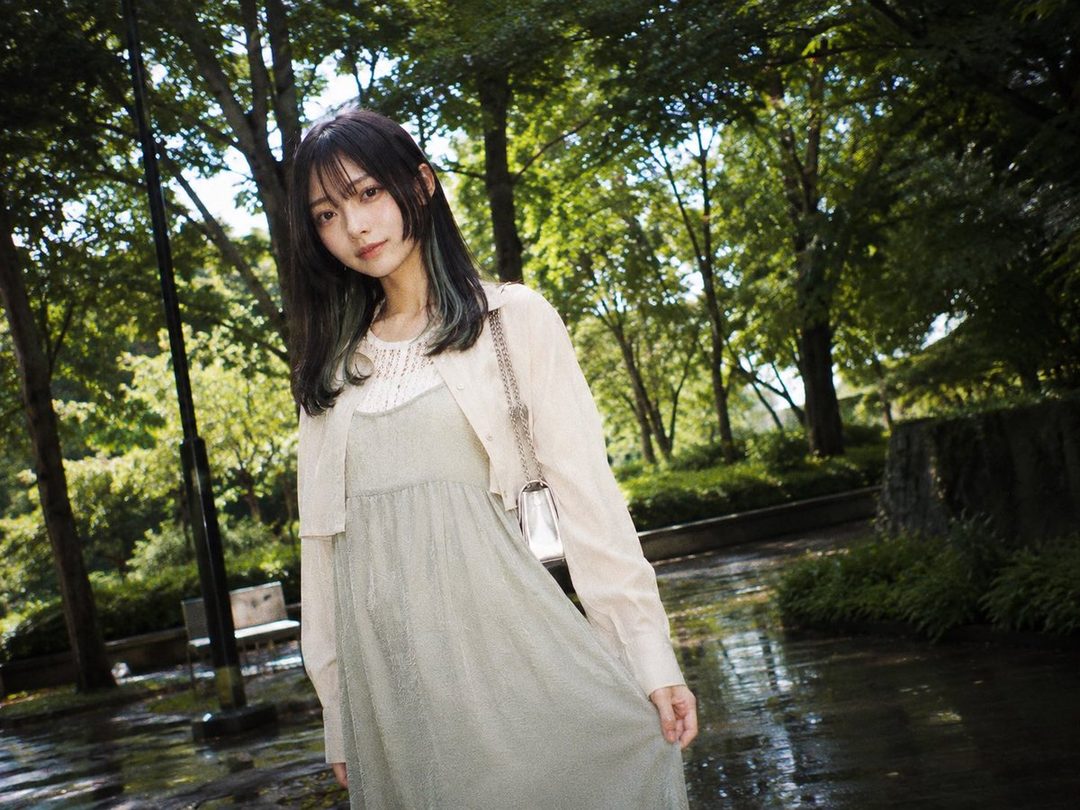 A young woman with long black hair and bangs, wearing a light green dress and a beige cardigan, stands in a park with lush green trees in the background.