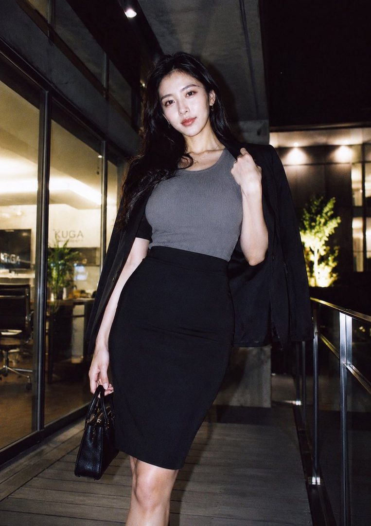 A woman in a grey top, black pencil skirt, and black blazer stands outside an office building at night, holding a black handbag.