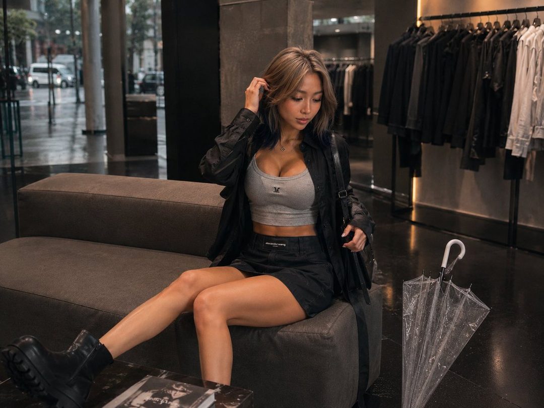 A stylish woman with blonde hair sits on a couch in a boutique, wearing a grey crop top, black mini skirt, and black boots, with an umbrella beside her.