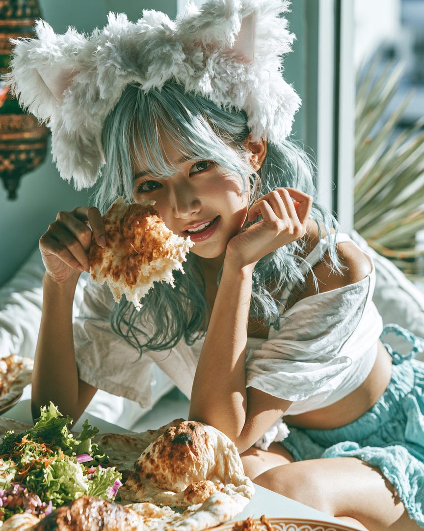 A young woman with light blue hair and fluffy cat ears is eating naan bread, with a salad and other food on the table.