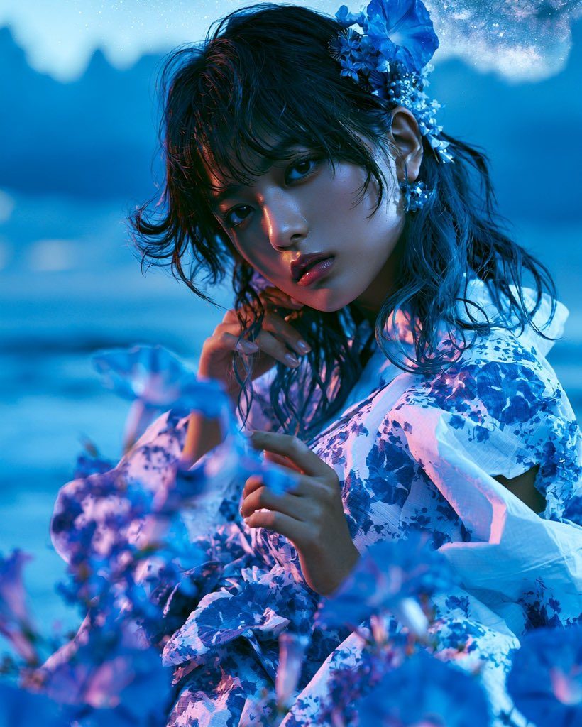 A young woman with dark hair adorned with blue flowers, wearing a blue and white floral yukata, poses outdoors amidst blue flowers with a dreamy blue and pink lighting.