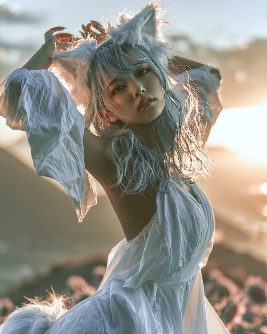 A beautiful AI model with light blue hair and cat ears, wearing a white flowing dress, poses outdoors during golden hour.