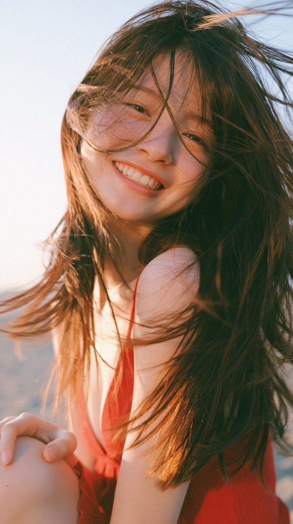 A young woman with long brown hair smiles brightly at the camera, her hair blowing in the wind on a sunny day at the beach.