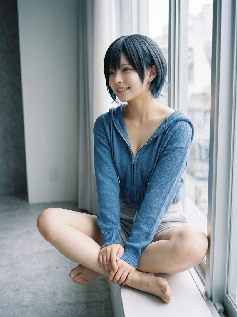 A young woman with short black hair sits cross-legged by a window, wearing a blue zip-up hoodie and grey shorts, looking to the side with a smile.