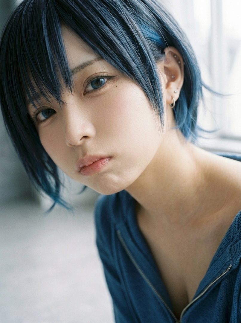 Close-up portrait of a young woman with short blue hair and blue eyes, wearing a blue hoodie.