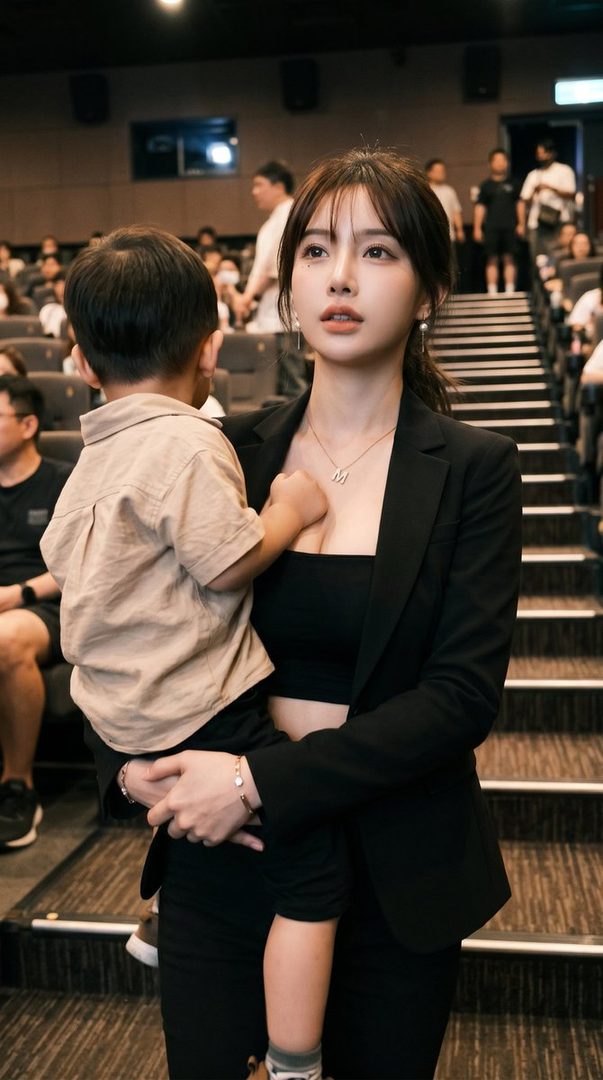 A woman in a black blazer and crop top holds a young child in a movie theater.