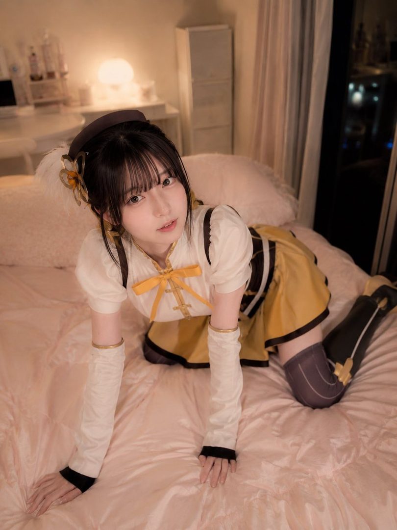 A young woman in a yellow and white outfit with a hat, posing on a bed indoors.