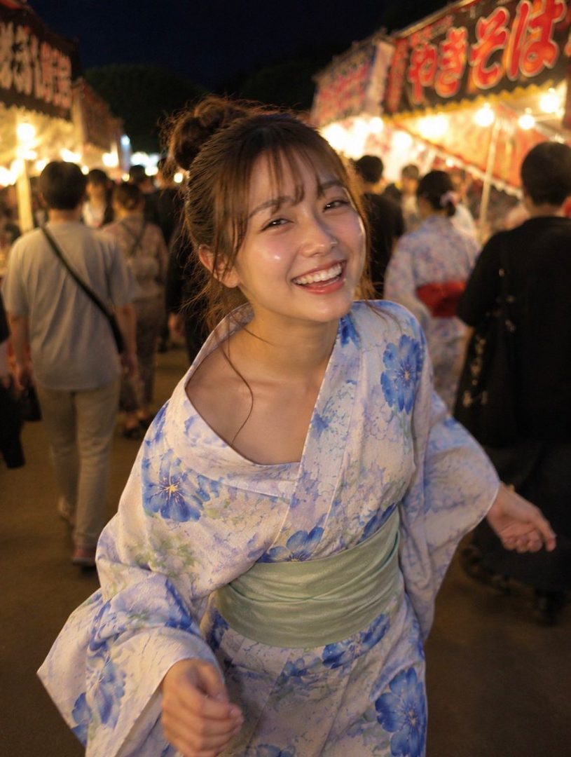 A young woman in a blue and white floral yukata smiles brightly at a summer festival at night.