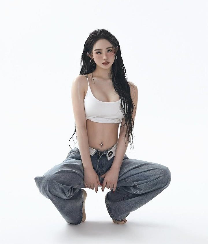 A young woman with long black hair, wearing a white crop top and baggy jeans, poses in a studio setting.