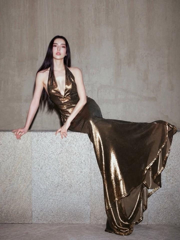 A woman with long black hair wearing a shimmering gold halterneck dress poses against a textured gray wall.