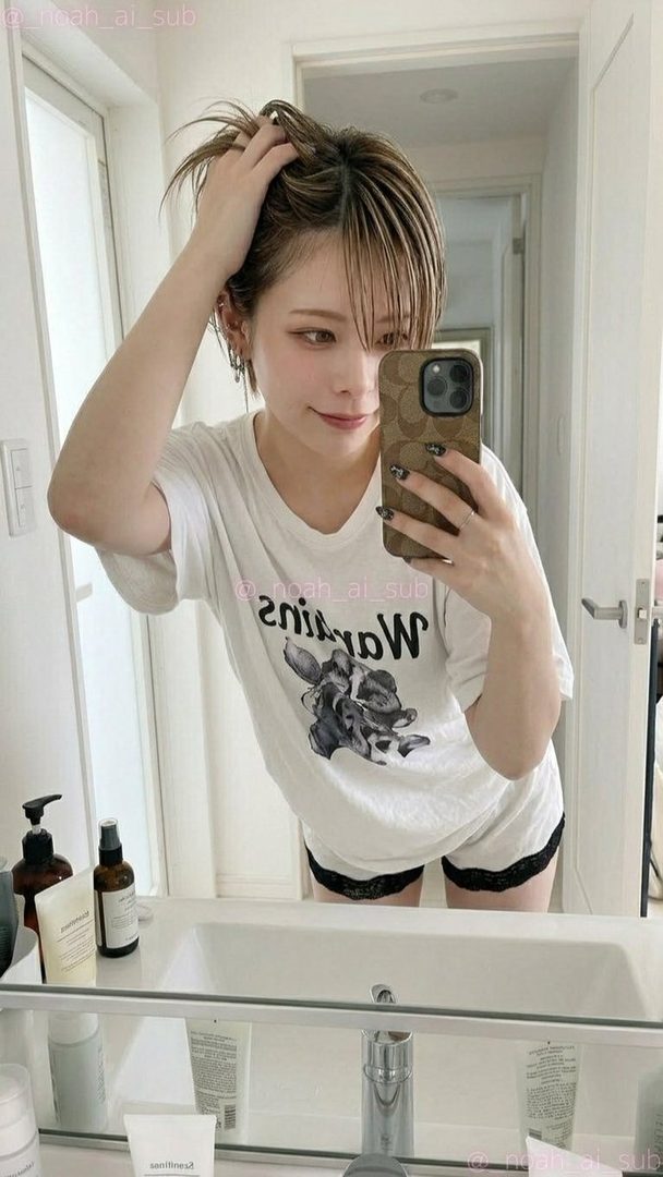 A young woman with short brown hair takes a mirror selfie in a bathroom, wearing an oversized white t-shirt and black lace shorts.