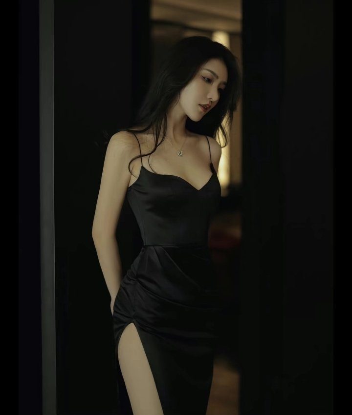 A beautiful East Asian woman with long black hair, wearing a black satin slip dress with a high slit, posing indoors.