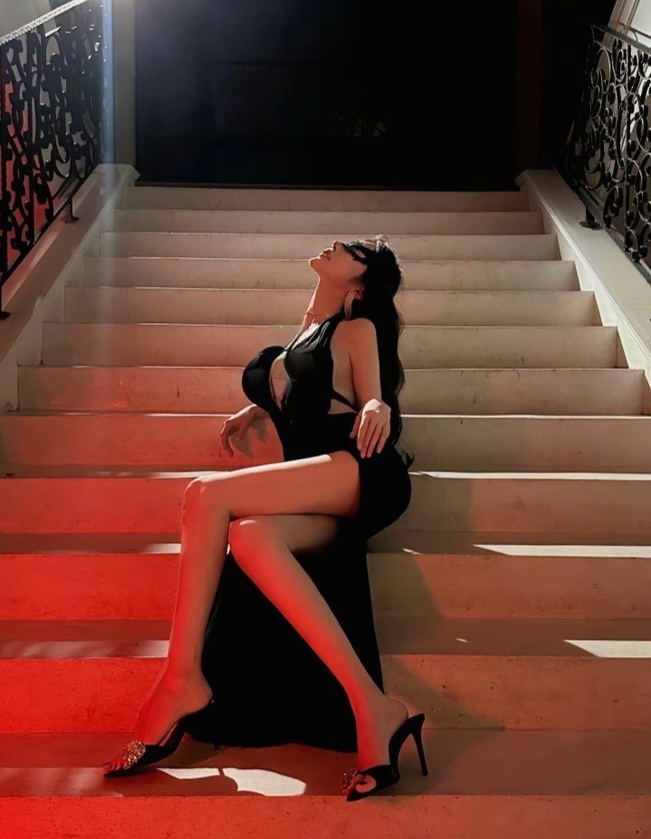 A woman in a black dress and mask sits on stairs, looking up.