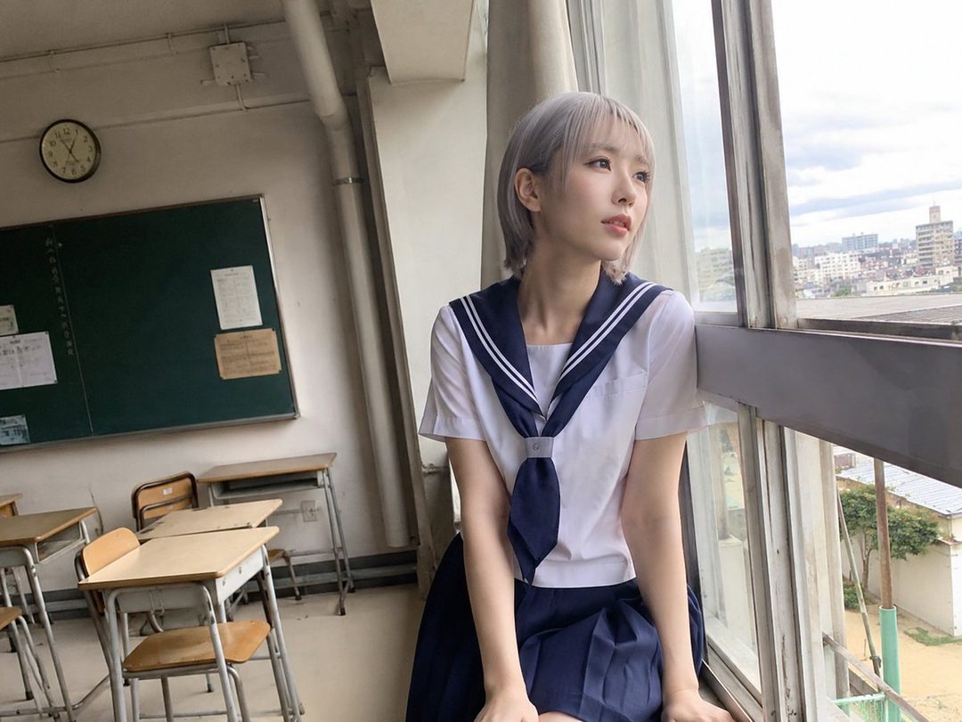 A young woman with short silver hair wearing a Japanese school uniform poses by a classroom window, looking out thoughtfully.