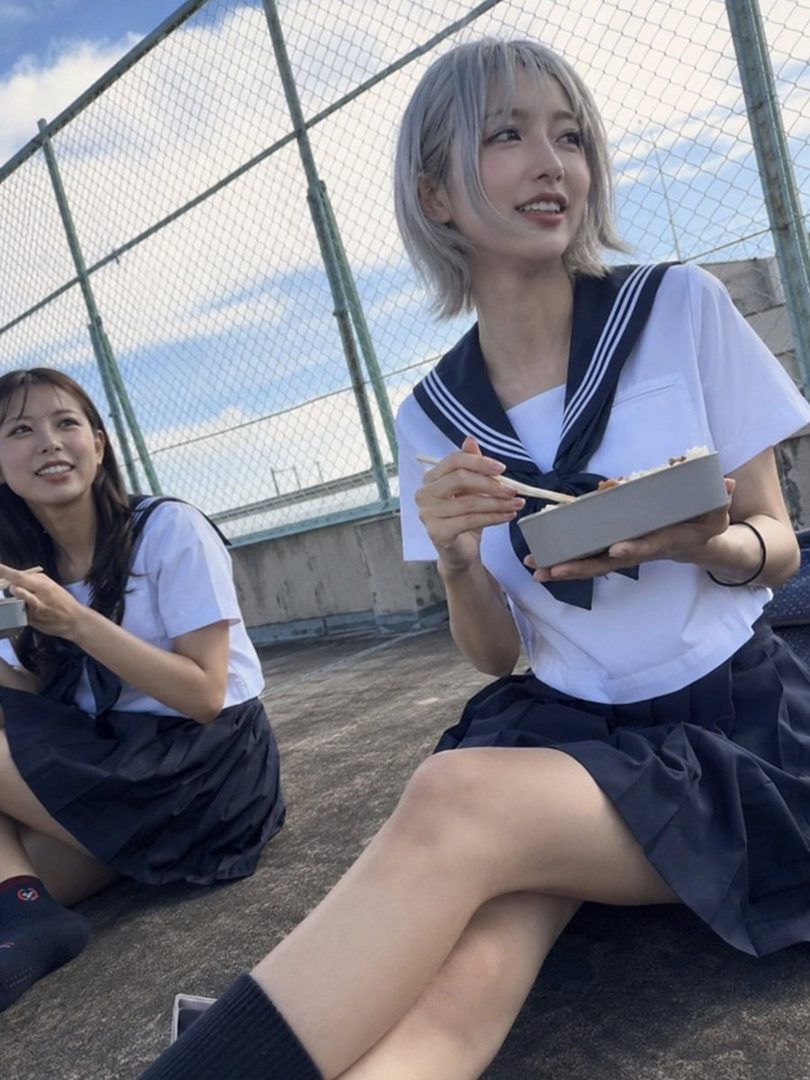 Two young women in school uniforms sitting on a rooftop, eating lunch.