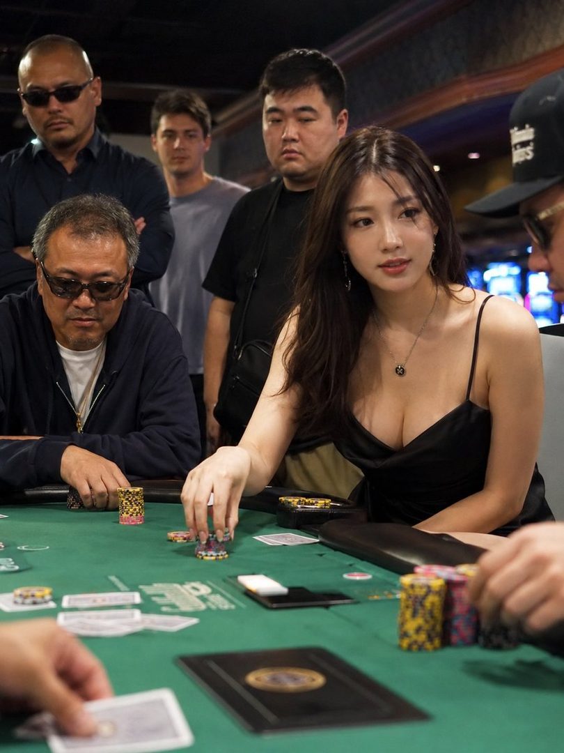 A beautiful East Asian woman in a black dress plays poker with several men watching.