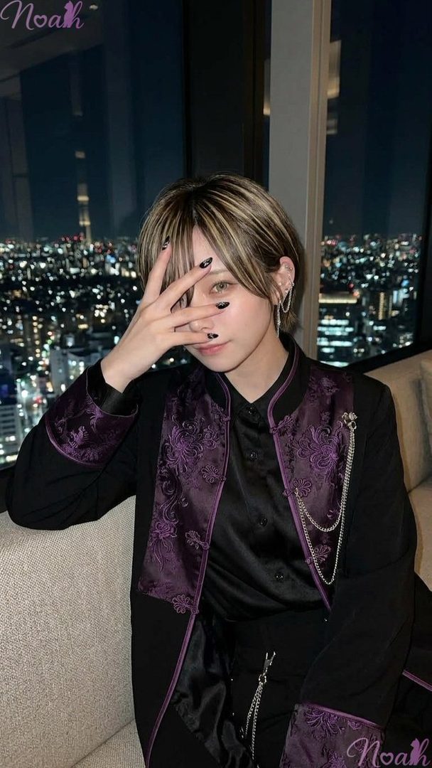 A person with short blonde-highlighted hair and black nail polish poses with their hand covering part of their face, wearing a black and purple jacket with chains, against a backdrop of a city night view.