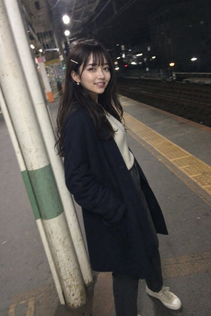 A young East Asian woman with long dark hair and bangs, wearing a white sweater and a dark coat, stands on a train platform at night, smiling at the camera.