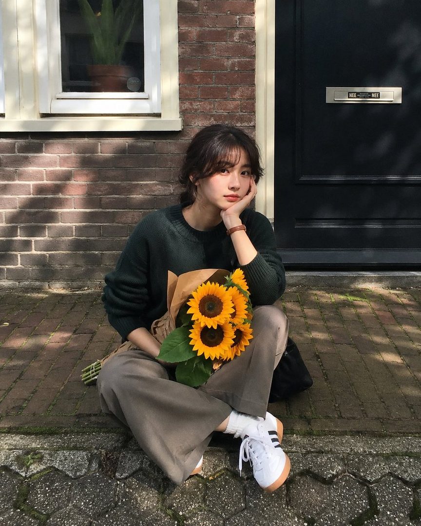 A young East Asian woman with dark hair sits on a sidewalk holding sunflowers, wearing a green sweater and grey pants.