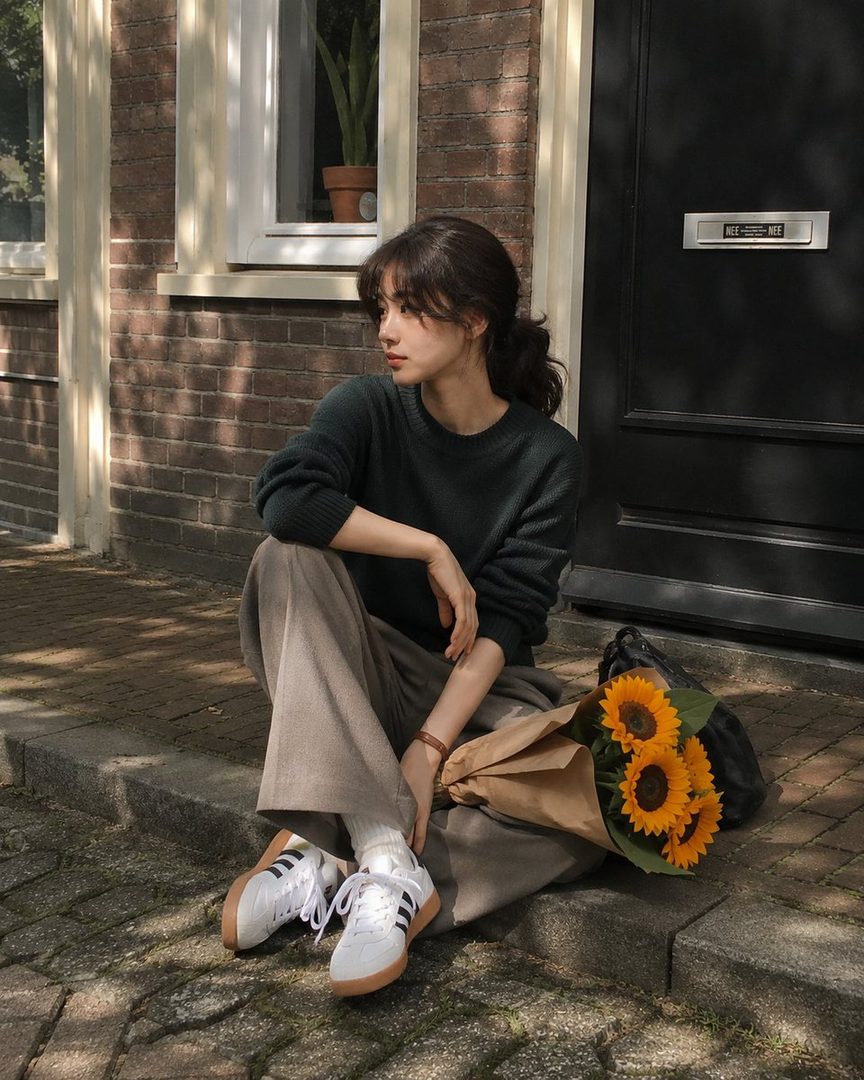 A young East Asian woman with dark hair in a ponytail sits on a sidewalk with a bouquet of sunflowers, wearing a green sweater and taupe pants.