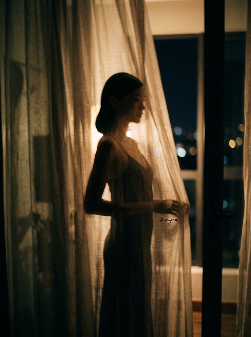 A woman in a slip dress stands by a window at night, partially obscured by curtains, with city lights visible in the background.