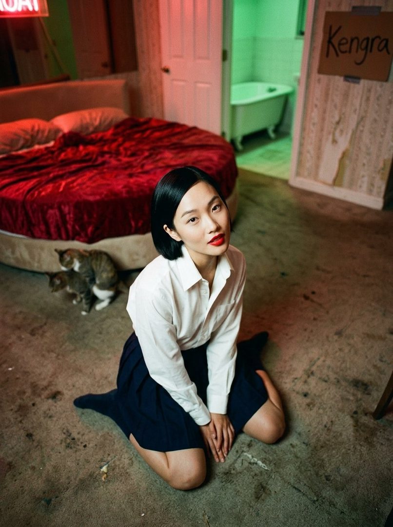 A young East Asian woman with short black hair and red lipstick kneels on the floor in a room with a large red bed and a bathtub visible in the background.