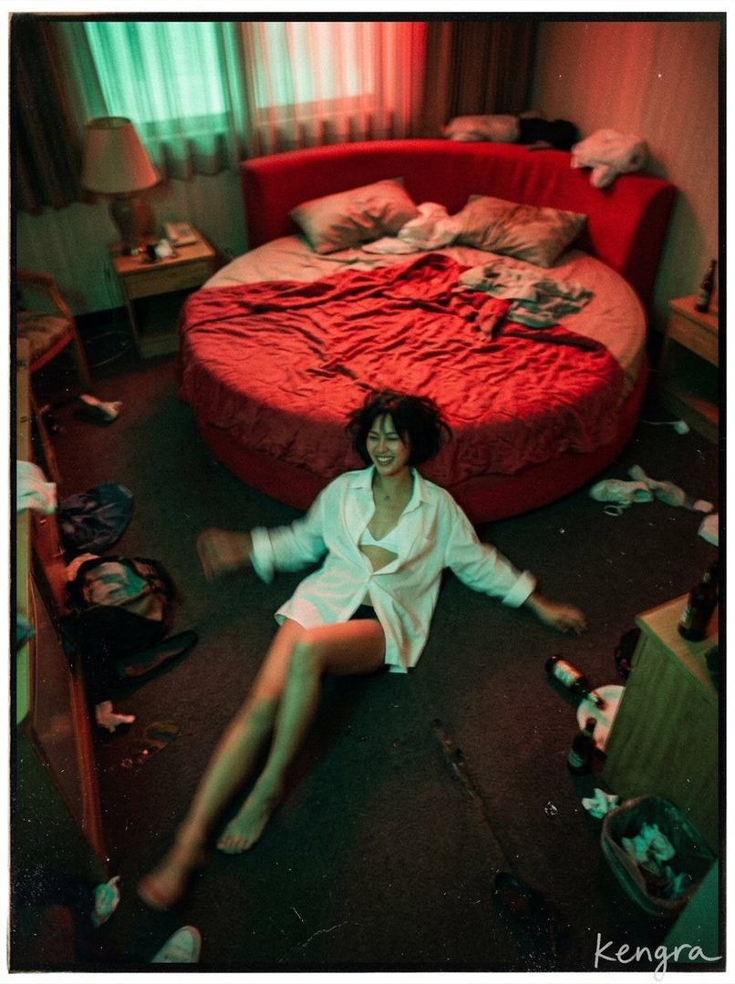 A young woman with short black hair smiles playfully while sitting on the floor of a messy bedroom, wearing an unbuttoned white shirt over a white bra.