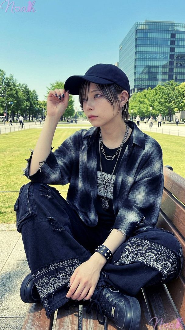A person with short black hair and highlights, wearing a plaid shirt, distressed jeans, and a baseball cap, sits on a park bench on a sunny day.