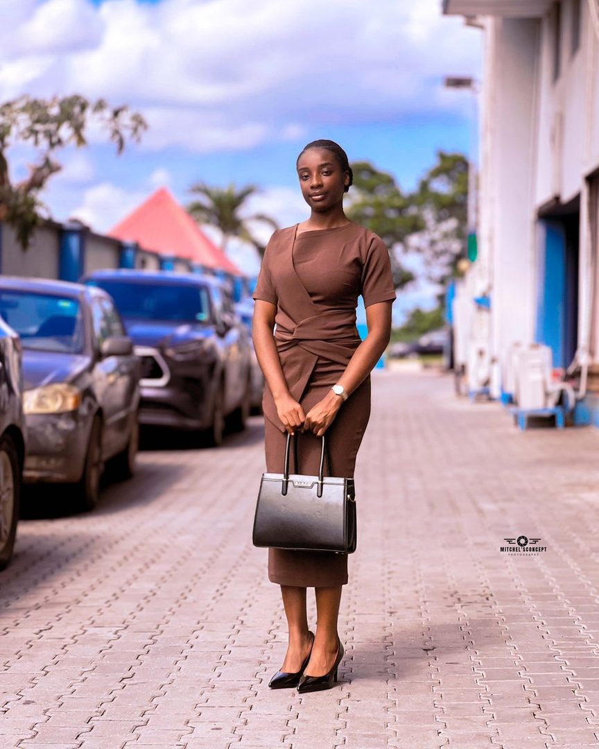 A young Black woman in a brown business dress and black heels stands outdoors holding a black handbag.