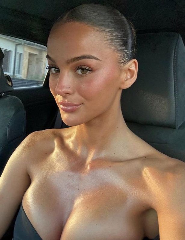 A young woman with her hair pulled back, wearing a strapless top, poses for a selfie in a car.