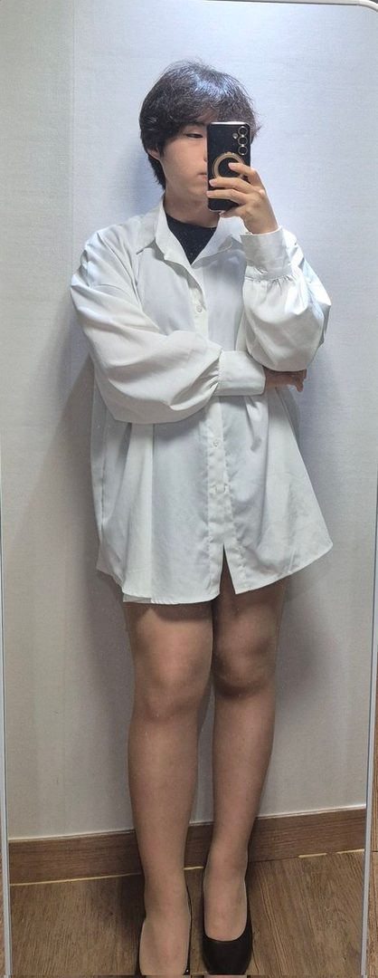 A person with short dark hair takes a mirror selfie, wearing an oversized white shirt and black flats, partially obscuring their face with a phone.