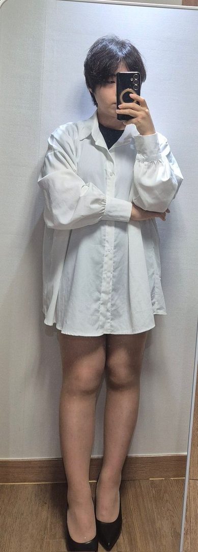 A person with short black hair takes a mirror selfie, wearing an oversized white button-up shirt and black heels.