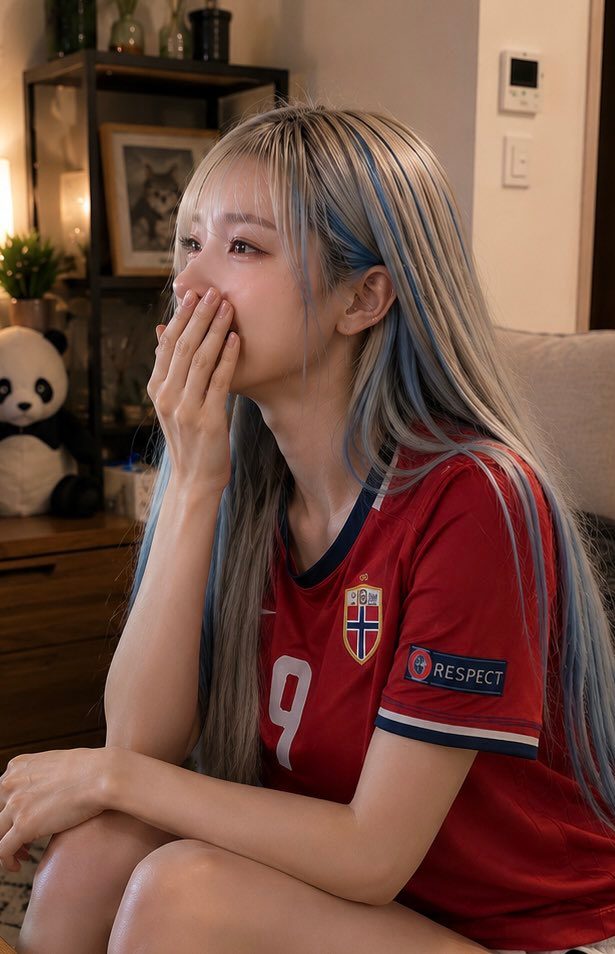 A young woman with long blonde hair with blue streaks, wearing a red Norway national football team jersey, covers her mouth with her hand and looks emotional.