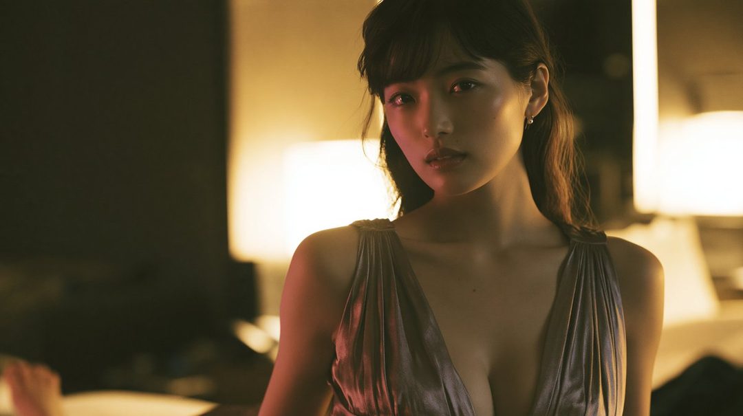 A beautiful East Asian woman with long brown hair, wearing a shimmering dress, poses elegantly indoors with soft lighting.