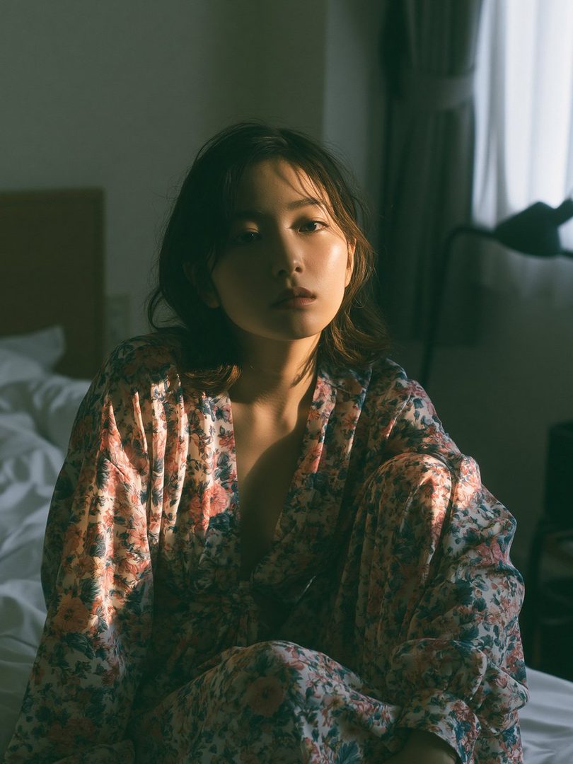 A young woman with shoulder-length brown hair sits on a bed, bathed in soft, golden sunlight streaming from a window. She wears a floral robe and looks directly at the camera with a serene expression.