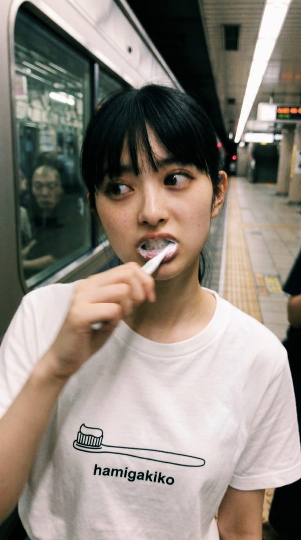 A young woman with black hair and bangs is brushing her teeth on a train platform, wearing a white t-shirt with a toothbrush graphic.