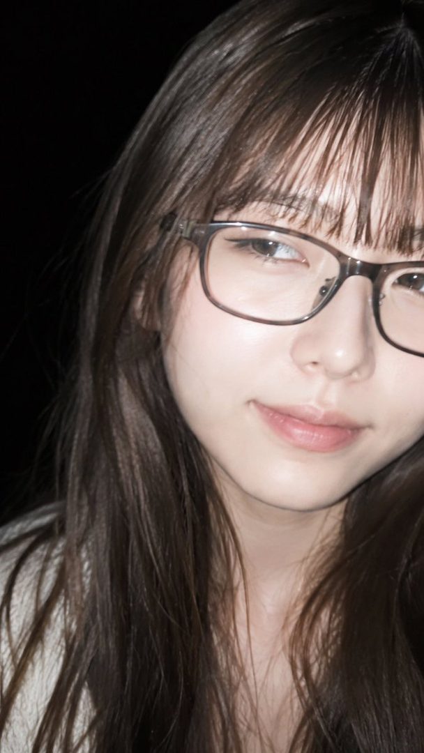 A close-up portrait of a young East Asian woman with long brown hair and bangs, wearing glasses, looking slightly to the side with a subtle smile.