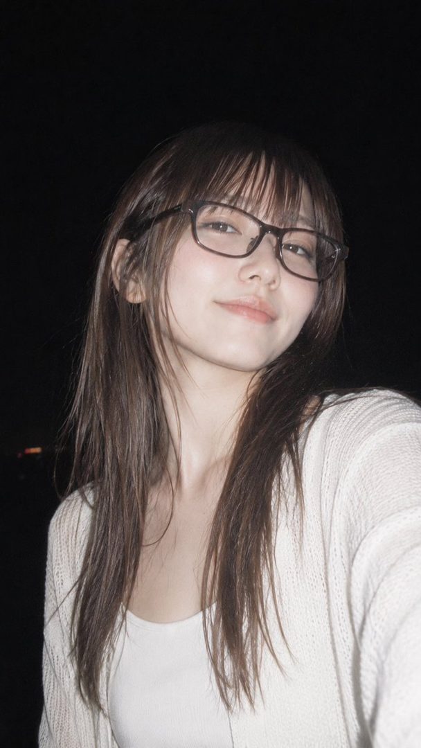 A young East Asian woman with long brown hair and bangs, wearing glasses and a white cardigan over a white tank top, smiles softly at the camera in a night outdoor setting.