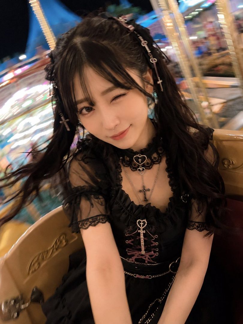 A young East Asian woman with black twintails and bangs winks at the camera, wearing a black gothic lolita dress and cross necklace, at a brightly lit amusement park at night.