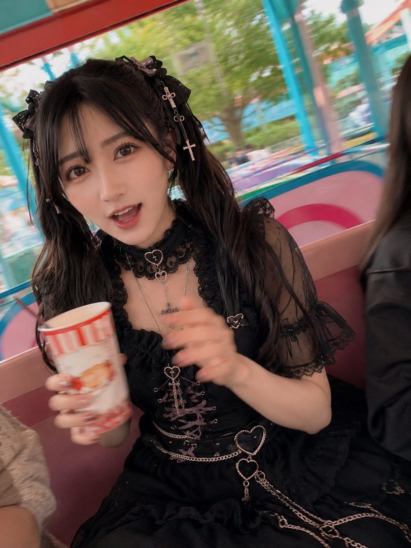 A young woman with black pigtails, wearing a black gothic lolita dress and holding a cup, smiles at the camera while riding a roller coaster at an amusement park.