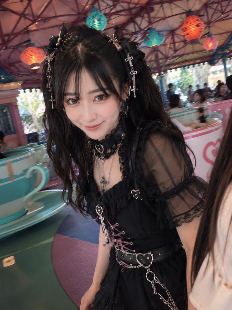 A young East Asian woman with long black hair and bangs, wearing a black gothic lolita dress and accessories, smiles at the camera in an amusement park.