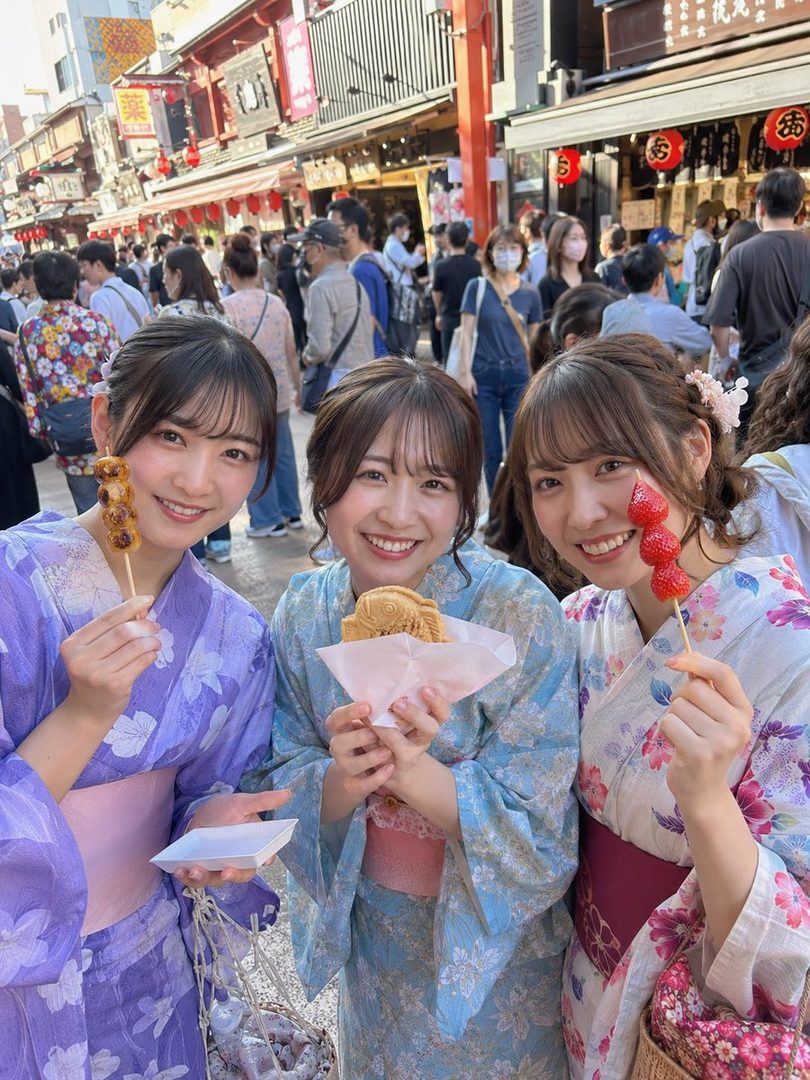 Three young Japanese women in colorful yukatas smile at the camera while holding traditional Japanese snacks on a busy street.