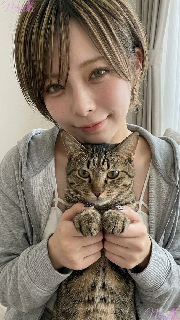 A young woman with short brown hair and green eyes smiles while holding a tabby cat close to her chest.