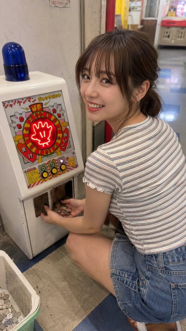 A young woman with brown hair tied in a ponytail, wearing a striped t-shirt and denim shorts, smiles at the camera while playing a coin-operated game machine.