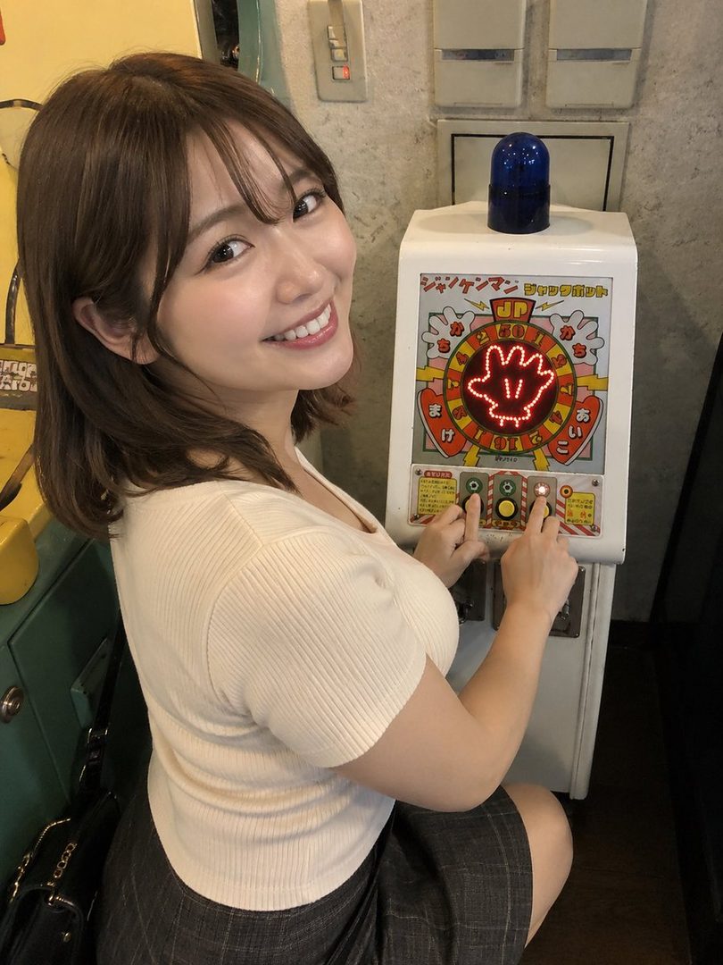 A smiling Japanese woman with short brown hair plays a retro arcade game indoors.