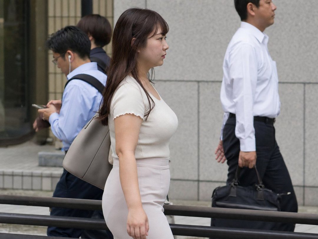 A woman in a light-colored top and skirt walks down a city street, seen from the side.